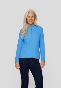 Hellblauer Pullover mit Stehkragen und halbem Reißverschluss, ausgestattet mit einer Fronttasche. Kombiniert mit dunkel gemusterten Hosen.