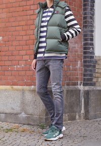 Zelená prošívaná vesta přes pruhované černobílé tričko s dlouhým rukávem, šedé džíny a zelené tenisky, na pozadí cihlové zdi.