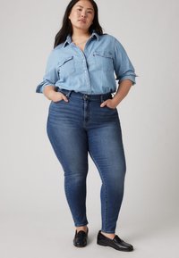 Camisa de ganga azul claro com botões e bolsos no peito, combinada com jeans skinny azul escuro. Modelo a usar sapatos mocassins pretos.