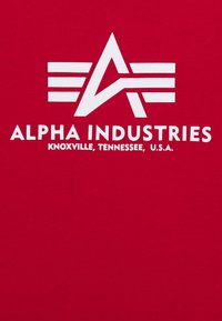 Camiseta de algodón roja con un gran logo blanco que presenta letras estilizadas "A" y rayas horizontales, junto con el texto "ALPHA INDUSTRIES".