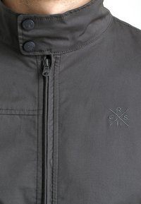 Szara kurtka zapinana na zamek z wysokim kołnierzem, z zatrzaskami, subtelnym logo haftowanym na piersi oraz gładką teksturą materiału.