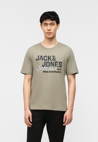 Jeune homme aux cheveux courts et bouclés portant un t-shirt vert olive clair avec le texte « JACK & JONES Superior New Aesthetics », debout devant un fond blanc.