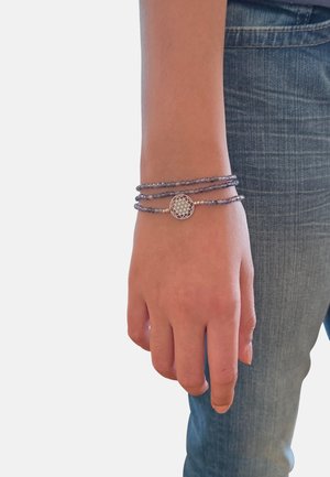 Mano e polso di una persona che indossa un braccialetto a tre fili di perle viola con un ciondolo geometrico circolare in argento, su sfondo di jeans e sfondo semplice.