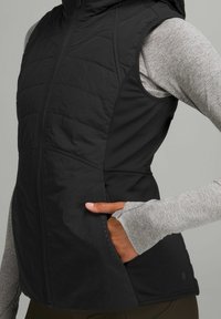 Gilet nero con cappuccio, con parte frontale trapuntata, tasche laterali e retro liscio. Abbinato a una maglietta a maniche lunghe grigia chiara con fori per il pollice.