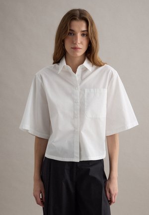 Junge Frau mit langen braunen Haaren, die ein weißes kurzärmeliges Hemd mit Knöpfen und schwarze Hosen vor einem schlichten beigen Hintergrund trägt.