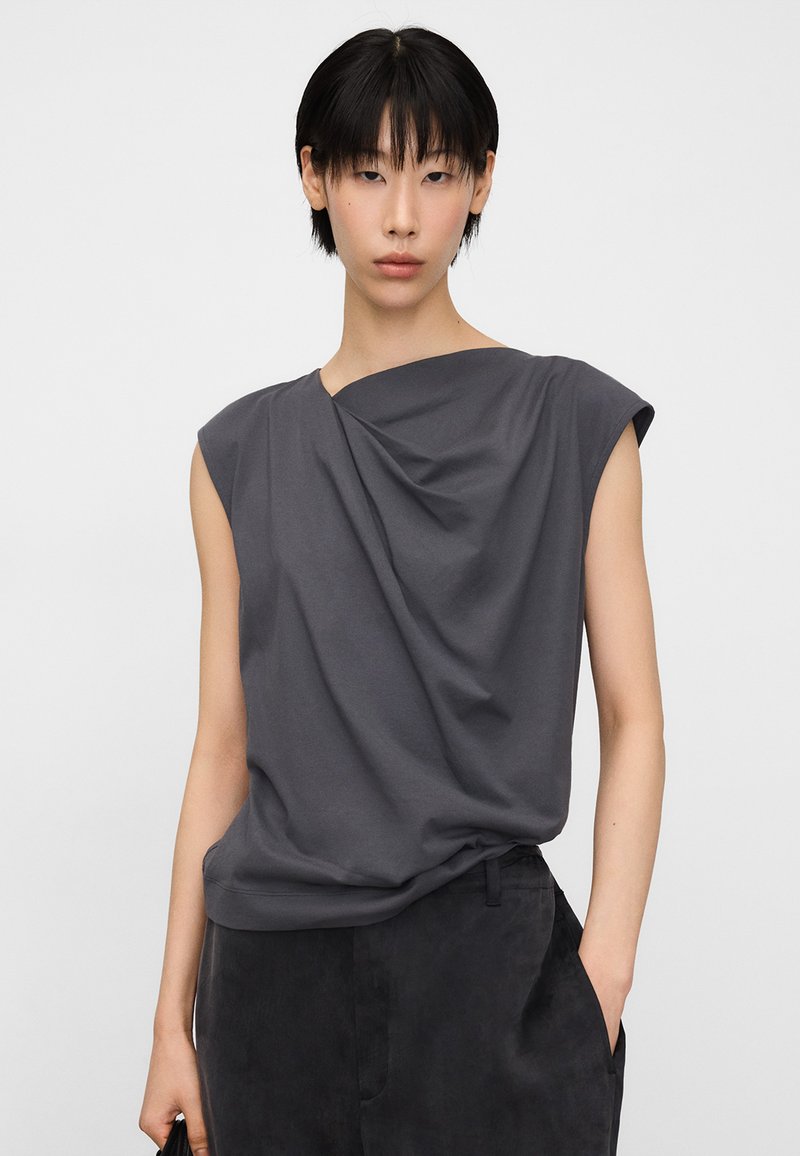 Person with short black hair wears sleeveless dark gray draped top and dark pants, standing with one hand in pocket against plain background.