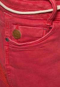 Pantalones vaqueros de mezclilla rojos que cuentan con un bolsillo delantero con un detalle de botón de bronce y un cordón blanco en la cintura. Acabado de tela texturizada.