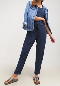 Femme portant une combinaison bleu marine, une veste en denim bleu clair drapée sur les épaules et des sandales noires, se tenant devant un fond neutre.