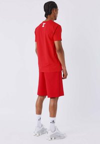 Homme de dos portant un t-shirt rouge et un short, des chaussettes blanches avec un logo, et des chaussures de sport blanches sur un fond uni.