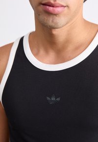 Svart ribbad linne med vit kant, med en liten tonad logotyp i mitten. Slät textur, figursydd design.