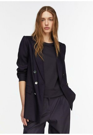 Frau mit langen, welligen Haaren, die einen dunkel zweireihigen Blazer über einem schwarzen Hemd und dunkler Hose trägt, steht vor einem weißen Hintergrund.