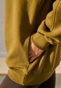 Olivengrøn sweatshirt med en struktureret overflade, der har en sidelomme og ribstrikkede ærmer. Stoffet viser små farveflekker for at give detalje.