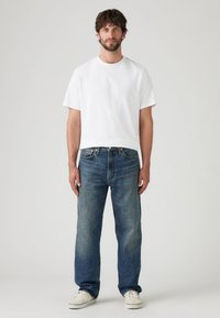 Bela majica s kratkimi rokavi v kombinaciji z modrimi kavbojkami s pravimi nogami. Kavbojke imajo posvetljen videz in so oblečene z belimi supergami.