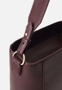 Sac en cuir bordeaux de forme rectangulaire, avec un accent en métal doré sur la sangle et des détails de couture précis. Texture lisse.