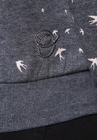Sweat-shirt gris en tissu texturé, présentant un motif subtil d'oiseaux blancs et un détail tressé près de l'ourlet.