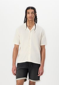 Short-sleeve, cream-colored button-up shirt with a textured knit pattern. Paired with dark denim shorts featuring a cuffed hem.