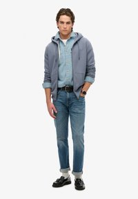 Lichtblauwe ritshoodie over een lichtdenim shirt, gecombineerd met opgerolde blauwe spijkerbroeken en zwarte instappers. Horloge zichtbaar om de pols.