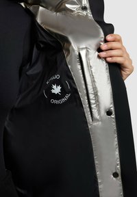 Melnā ārējā veste ar spīdīgu sudraba odere, kurā ir redzama roka, kas to tur vaļā. Uz vests ir logo ar kļavas lapu, ar uzrakstu "KHUJO ORIGINAL."