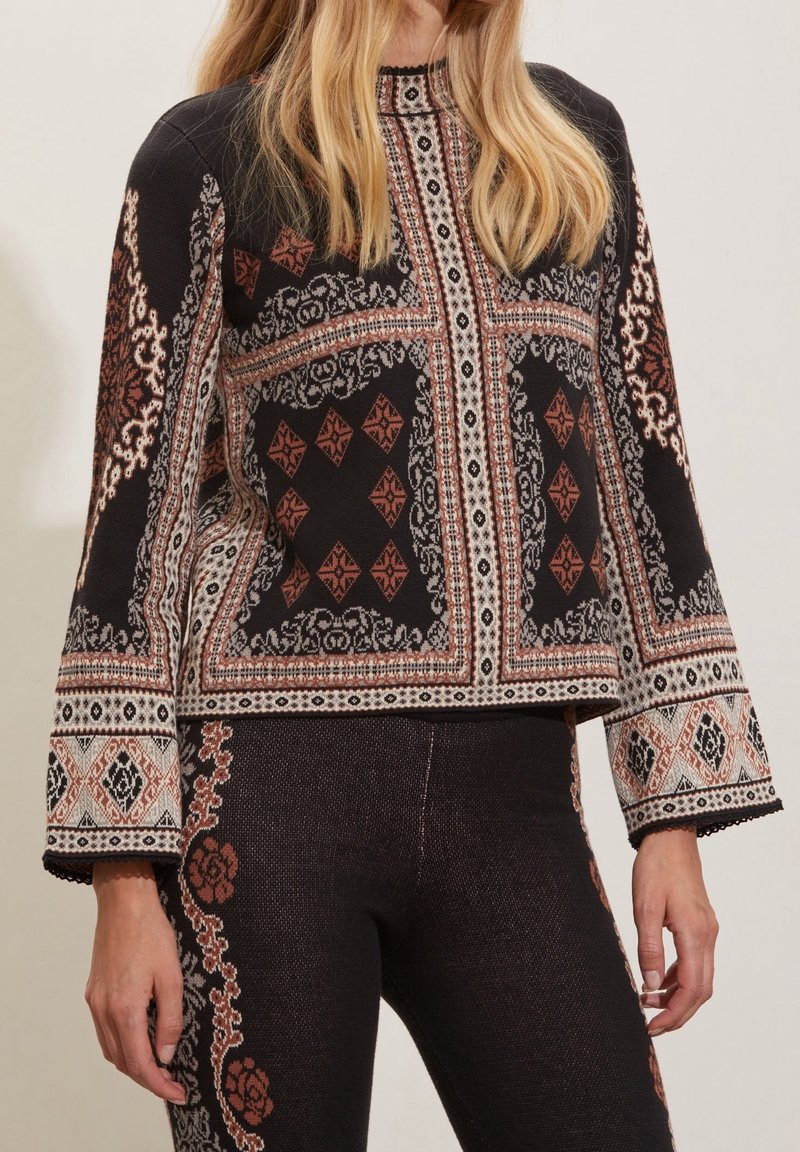 Femme porte un haut en maille noir et un pantalon aux motifs géométriques et floraux symétriques dans des tons marron, blanc et beige.