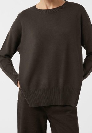 Person trägt dunkelbraunen Langarm-Strickpullover und passende Hose, steht mit den Händen in den Taschen vor einem einfarbigen Hintergrund.