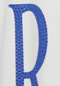 Tissu blanc avec une grande lettre bleue décorée de strass brillants en relief. La lettre est en gras et caractérisée par une texture lisse.