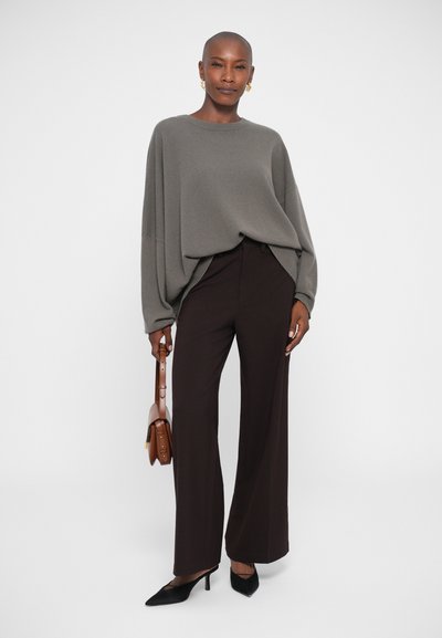 Szürke, oversized, ejtett vállú pulóver, sötétbarna, széles szárú nadrággal párosítva. A modellen egy kis barna kézitáska van, és fekete magassarkú cipőt visel.