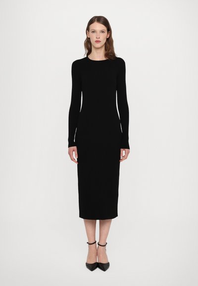 Robe en tricot côtelé noire avec manches longues, silhouette ajustée et ourlet longueur genou. Associée à des escarpins noirs à talons pointus et à brides de cheville.
