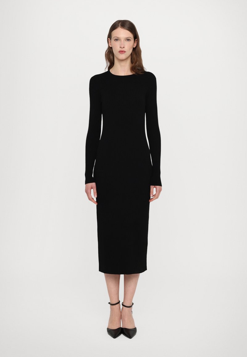 Vestido negro de punto acanalado con mangas largas, silueta ajustada y dobladillo a la altura de la rodilla. Combinado con tacones negros de punta y correas en el tobillo.