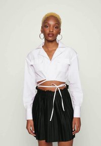 Mulher com cabelo loiro curto, vestindo uma blusa branca de manga curta com atilhos e uma saia preta plissada, de pé contra um fundo claro.