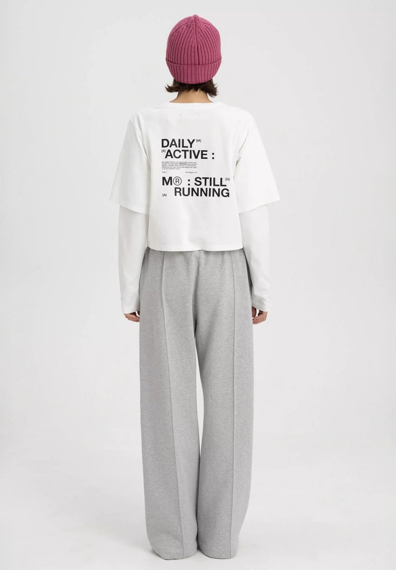 Witte cropped t-shirt met zwarte tekst op de achterkant. Gecombineerd met grijze wijde broek en een roze geribbelde beanie.