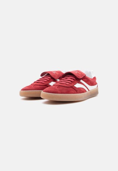 Baskets en daim rouge avec des bandes blanches, des semelles en gomme, des lacets, et une languette repliée sur la languette, posées sur un fond blanc.
