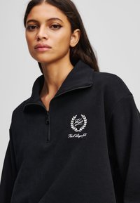 Čierna mikina na zips s vysokým golierom, vyšitým logom s vavrínovým dizajnom na ľavej hrudi a jemnou textúrou.