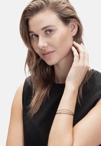 Tunn metallarmband med tre trådar i silver och svart, bärs på ett naket handled, mot en neutral bakgrund.