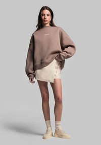Felpa marrone oversize con polsini a coste, gonna avvolgente color crema con dettagli a sorpresa, e sneakers chunky beige. Texture lisce, vestibilità comoda.