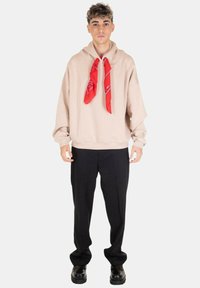 Felpa oversize beige con cappuccio e cordino, con una sciarpa rossa a motivo al collo, abbinata a pantaloni neri e scarpe nere lucide.