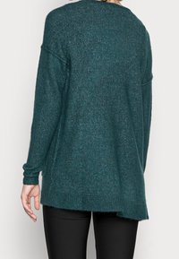 Maglione turchese lavorato a maglia con maniche lunghe e scollatura arrotondata, caratterizzato da un orlo posteriore leggermente più lungo e una texture morbida.