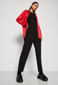 Rød satengskjorte, svart tettsittende topp, høyt liv svart bukse, skinnende svarte loafers med dekor. Enkel bakgrunn, modell som poserer selvsikkert.