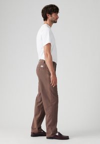 Bruine katoenen broek met een relaxte pasvorm, heeft zijzakken en een achterzak, gecombineerd met een wit T-shirt en donkere instappers.