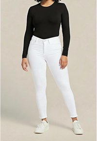 Top negro de manga larga combinado con vaqueros blancos ajustados. Completa el conjunto un par de zapatillas blancas con cordones. El fondo es de un tono neutro.