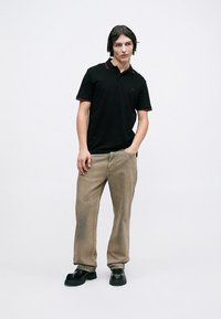 Young man standing with one hand in pocket, wearing black polo shirt, beige loose pants, and black chunky shoes against plain light background.