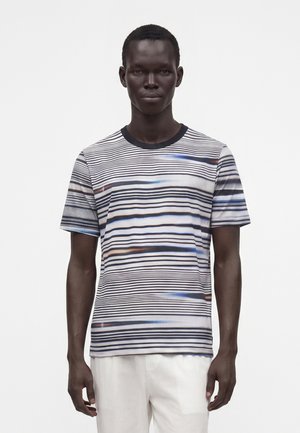Homme portant une chemise à manches courtes avec de fines rayures horizontales noires et bleues et un pantalon blanc, debout devant un fond blanc uni.