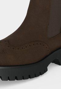 Botte en daim marron avec panneau latéral élastique, détails brogue sur la tige et semelle épaisse en caoutchouc noir cranté.