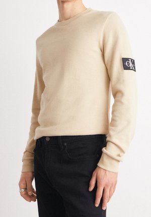 Un homme porte un pull beige à manches longues avec un écusson logo CK sur le bras et un jean noir, debout devant un fond clair et uni.
