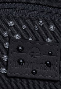 Sac en tissu noir avec des gouttes d'eau sur la surface, avec un patch en cuir noir texturé embossé portant "JOHNNY URBAN".