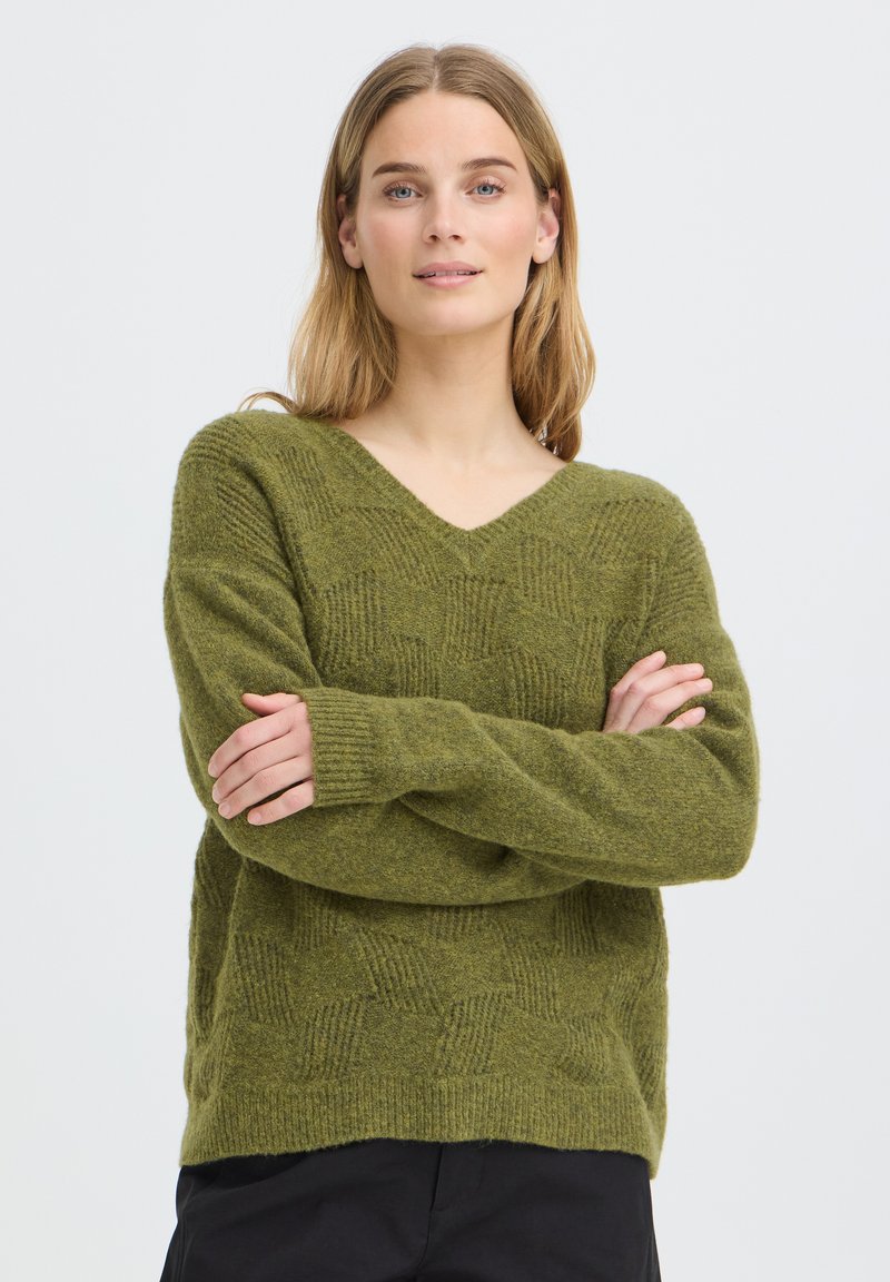 Jeune femme aux cheveux blonds portant un pull vert texturé à col en V et un pantalon noir, debout les bras croisés devant un fond uni.
