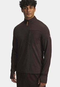 Jonge man in een zwart langarmoenen kwart-zip top met een zak aan de voorkant en duimhole tegen een effen achtergrond.