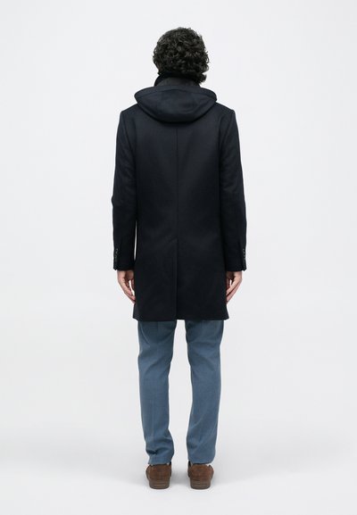 Cappotto scuro di lana con cappuccio, linea sagomata e spacco singolo sul retro; indossato sopra pantaloni grigio chiaro e scarpe marroni. Texture liscia.