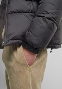 Sort nedjakke med kviltet tekstur og snørede ærmer; beige bukser med ternet mønster og brede lommeskæringer.