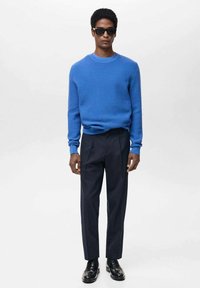 Blauwe geribde trui, marineblauwe plooibroek, zwarte schoenen, model met zonnebril, gladde texturen, moderne snit, strakke lijnen.