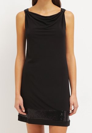 Schwarzes ärmelloses Kleid mit drapiertem Ausschnitt und einem paillettenbesetzten Band nahe dem Saum, getragen von einer stehenden Person vor schlichtem Hintergrund.